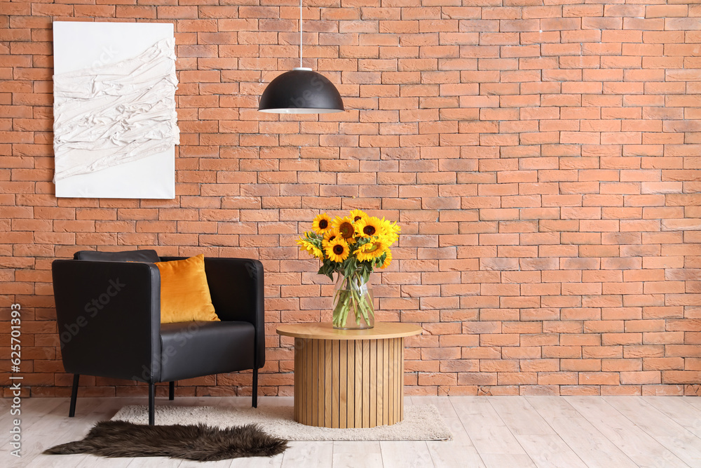 Vase with beautiful sunflowers on coffee table and black armchair near brown brick wall