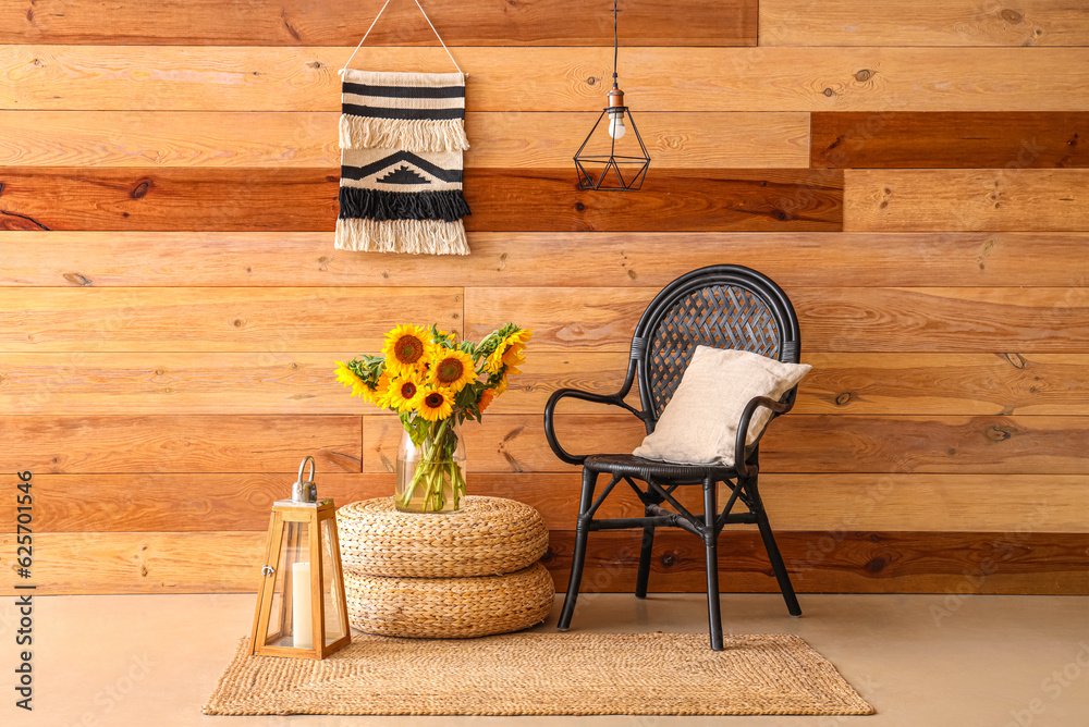 Vase with beautiful sunflowers on wicker pouf and armchair near wooden wall