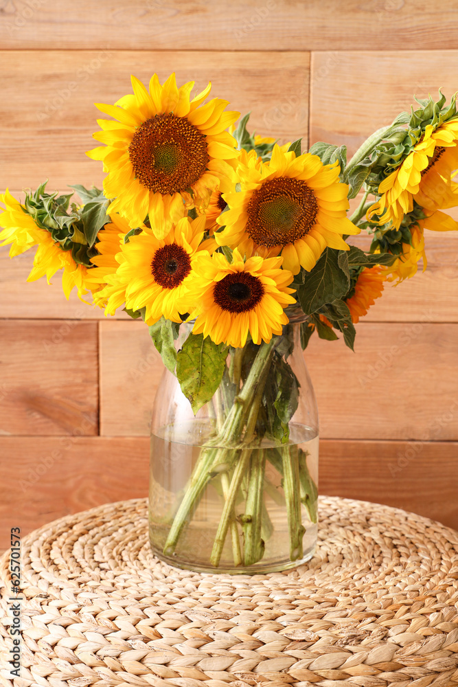 Vase with beautiful sunflowers on wicker pouf near wooden wall, closeup