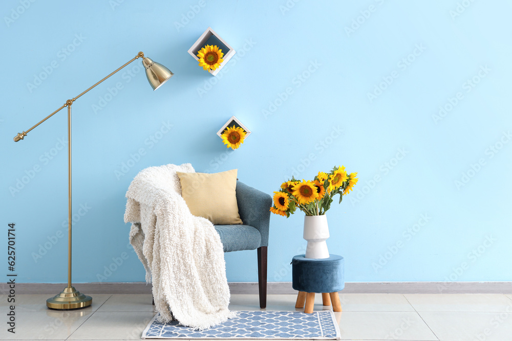 Vase with beautiful sunflowers on pouf and cozy armchair near blue wall