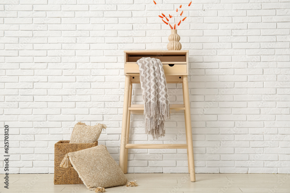 Table with cozy blanket and cushions in wicker basket near white brick wall