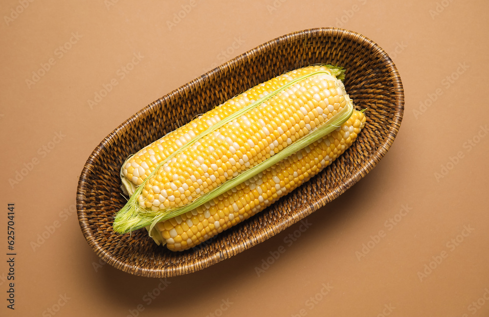 Wicker bowl with fresh corn cobs on brown background