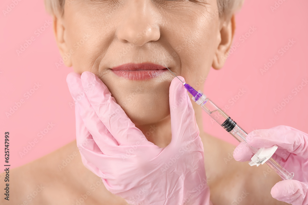 Mature woman receiving lip filler injection on pink background, closeup