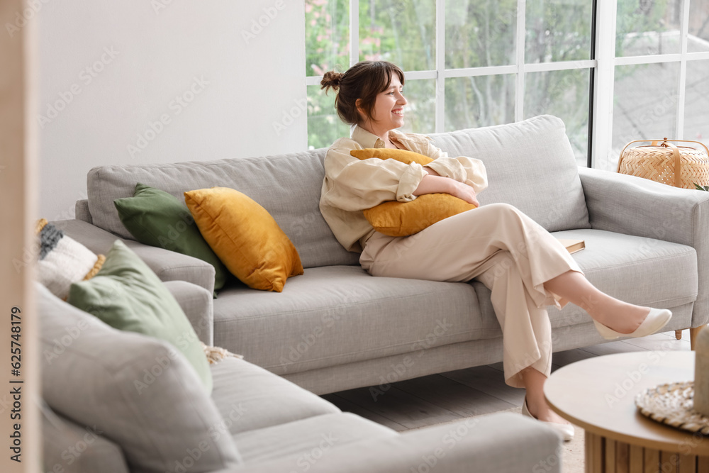 Happy young woman sitting on grey sofa in interior of light living room