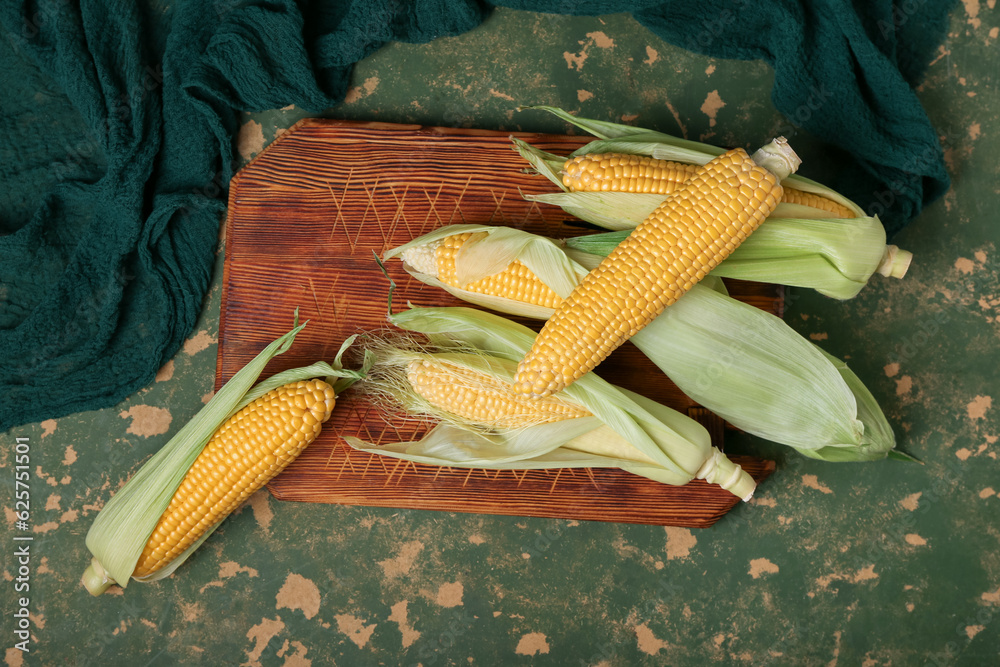 Wooden board with fresh corn cobs on green background