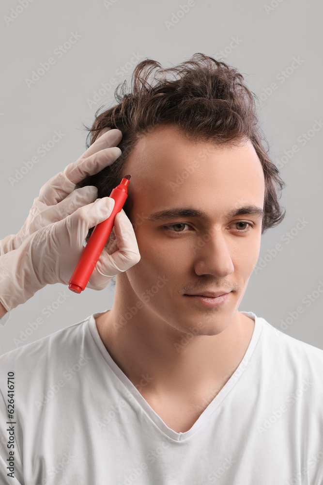 Doctor marking young mans forehead with hair loss problem on grey background, closeup