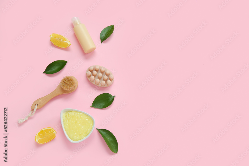 Bowl of lemon body scrub with cosmetic bottle, massage soap bar and brush on pink background