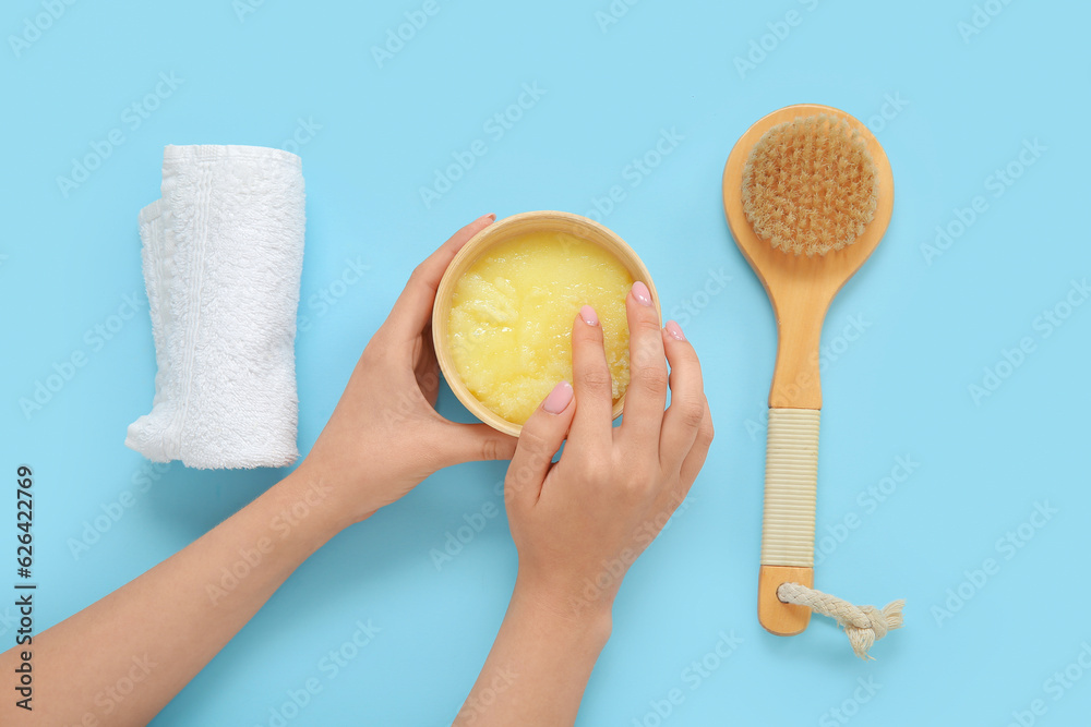 Female hands with bowl of lemon body scrub, towel and massage brush on blue background