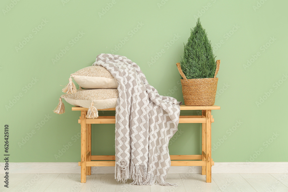 Wooden bench with blanket, cushions and plant in wicker basket near green wall