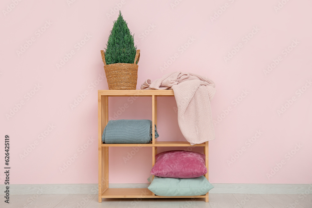 Shelving unit with cozy blankets, cushions and houseplant near pink wall