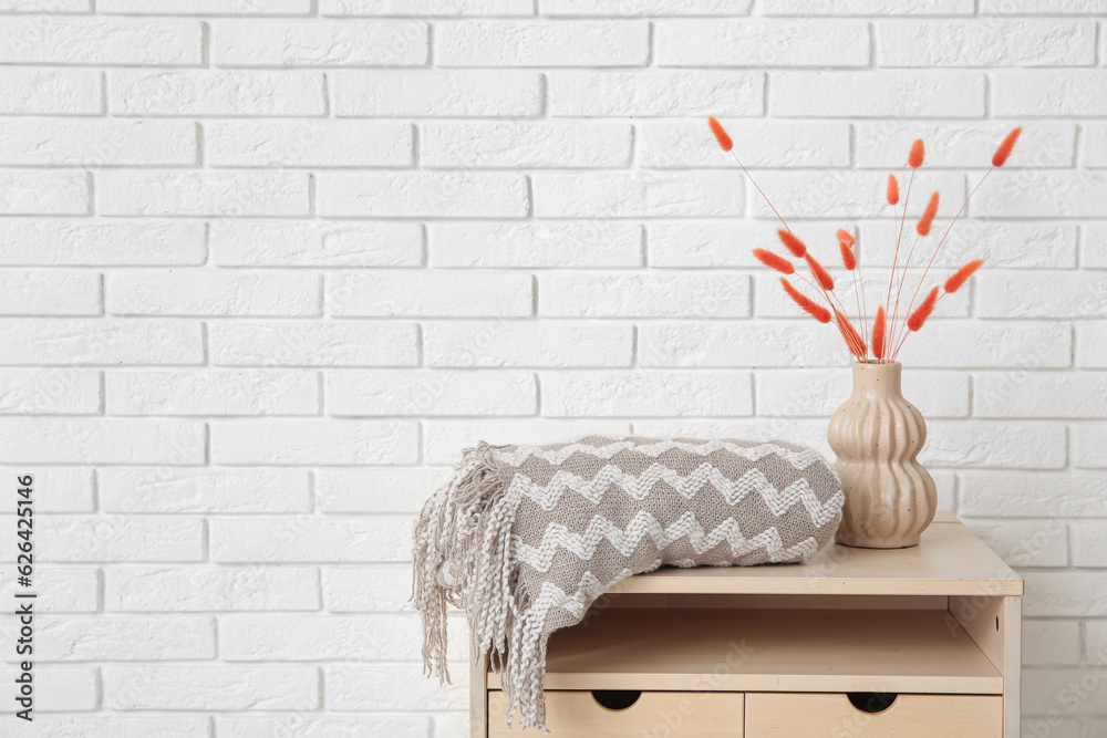 Table with cozy blanket and dried flowers in vase near white brick wall