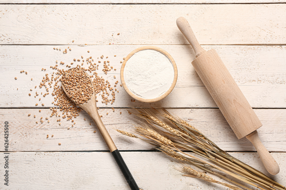 Bowl of wheat flour and spoon with grains on white wooden background