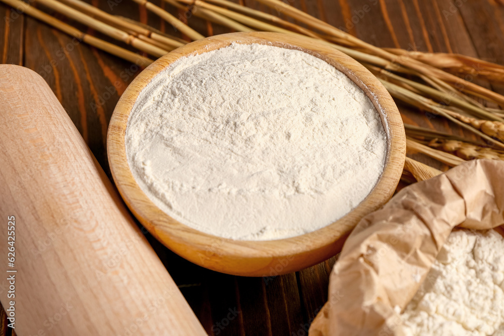 Paper bag and bowl of flour with wheat ears on wooden background