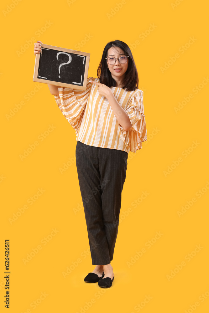 Female Asian teacher pointing at chalkboard with question mark on yellow background