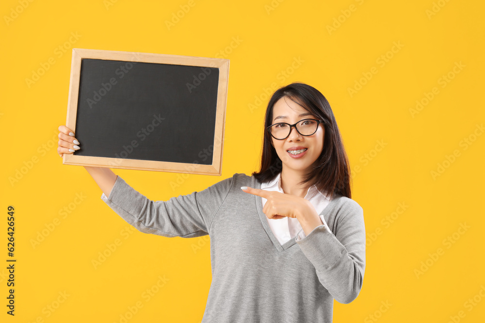 Female Asian teacher pointing at chalkboard on yellow background