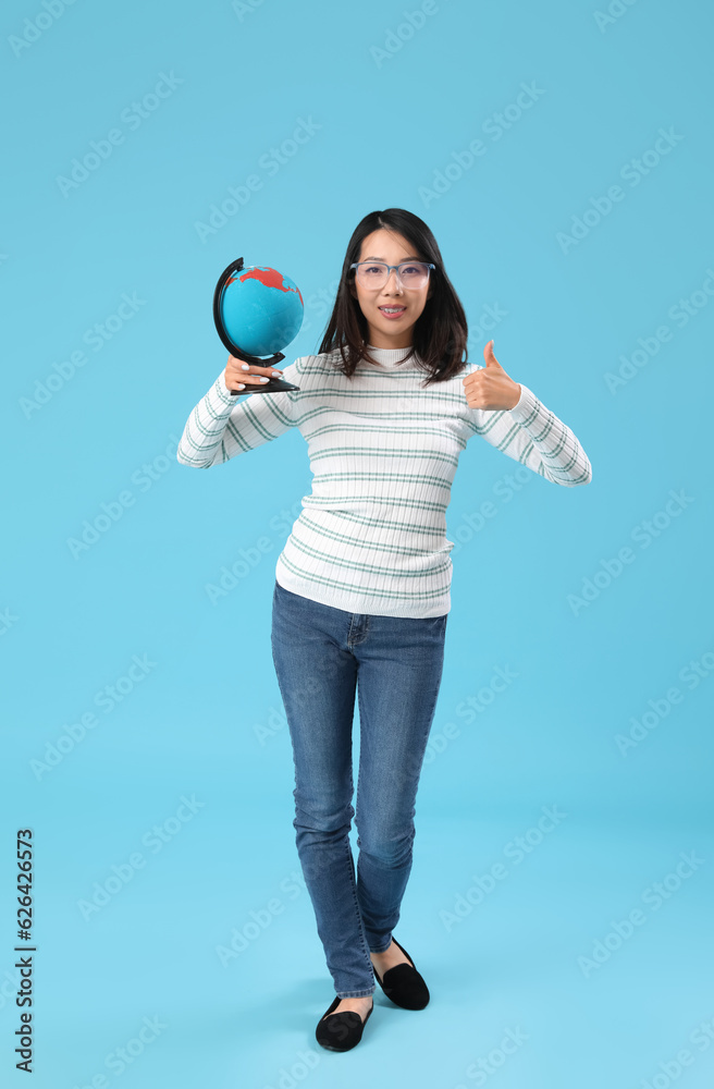 Asian Geography teacher with globe showing thumb-up on blue background