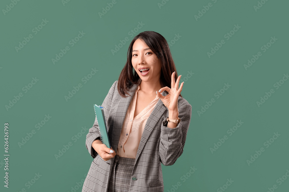 Female Asian teacher with notebook showing OK on green background