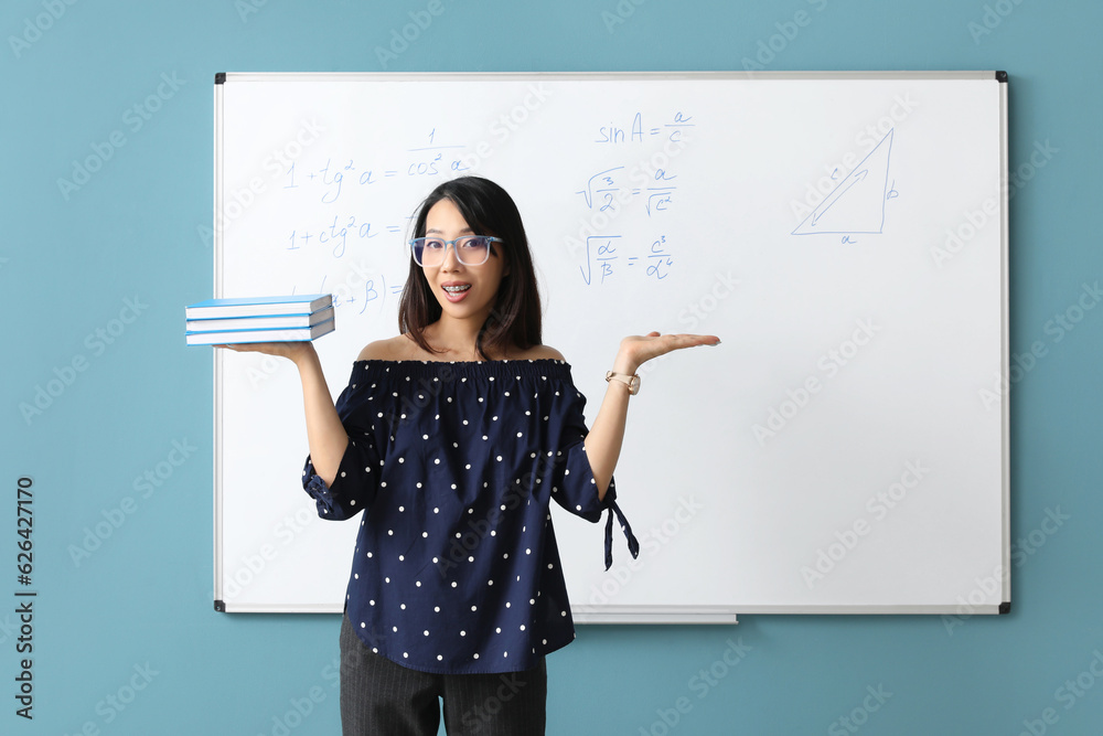 Asian Math teacher with books near flipboard in classroom