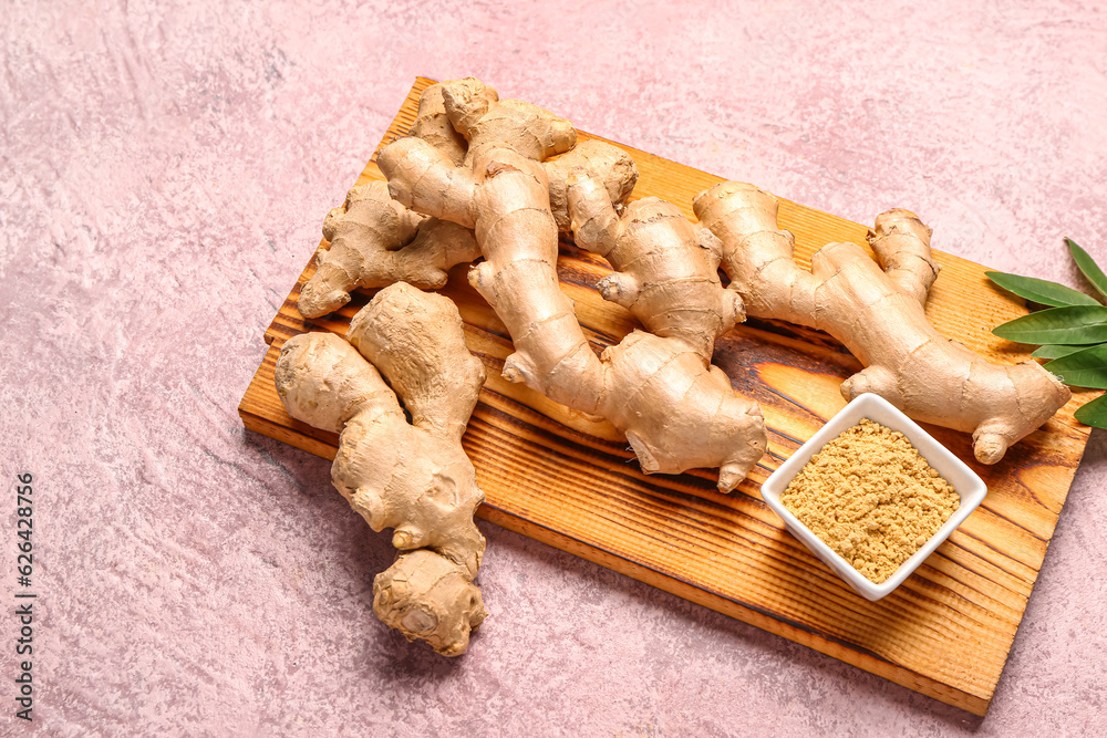 Wooden board with fresh ginger roots and bowl with dried powder on pink background