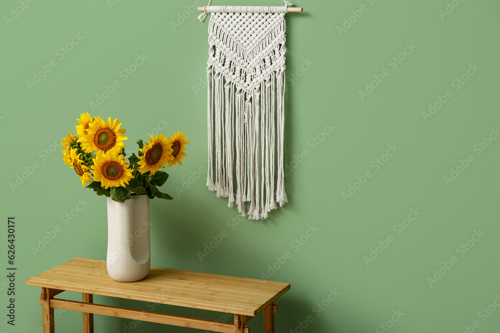 Vase with beautiful sunflowers on table and decor hanging on green wall in room