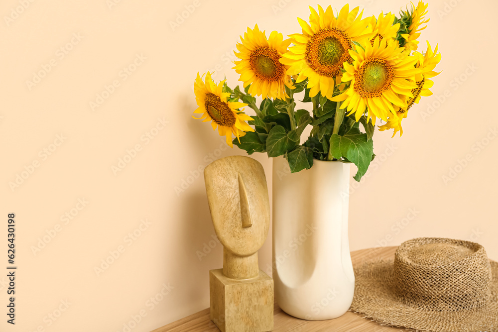 Vase with beautiful sunflowers and hat on table near beige wall in room, closeup