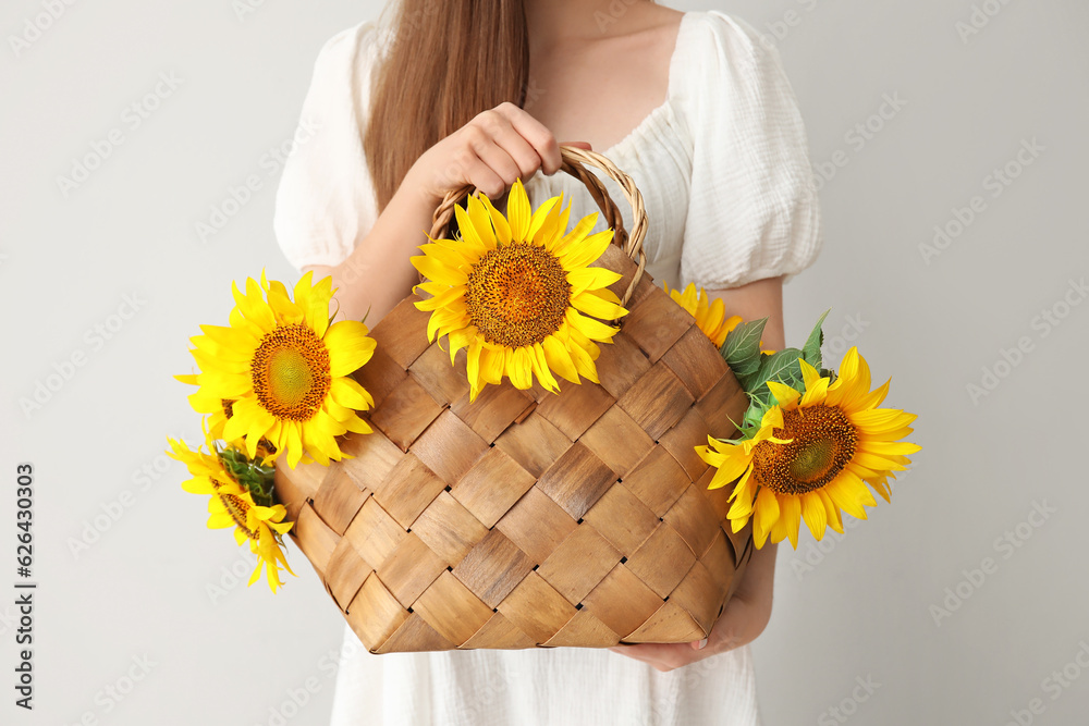 Young woman with beautiful sunflowers in wicker basket on light background