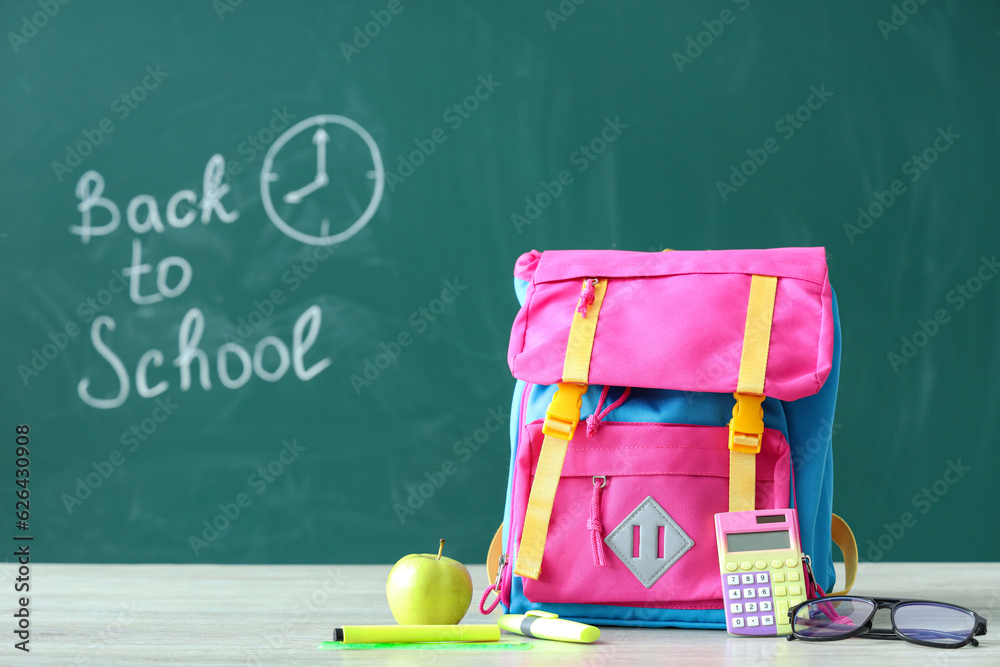 Backpack and supplies on table near green chalkboard with text BACK TO SCHOOL