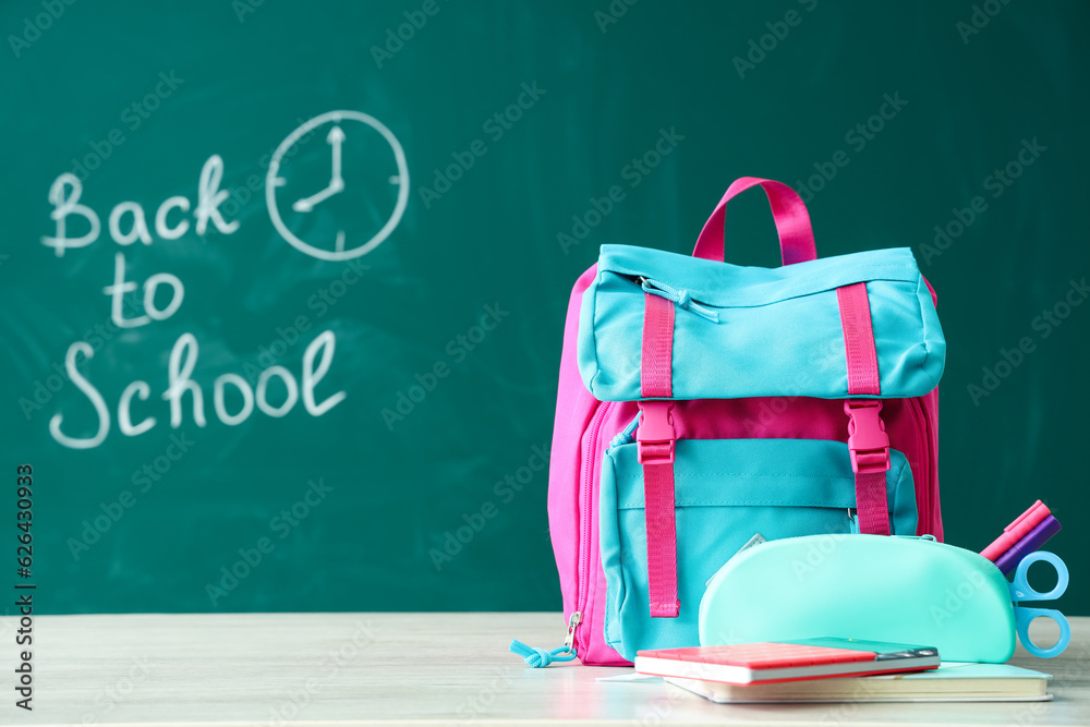 Backpack and supplies on table near green chalkboard with text BACK TO SCHOOL