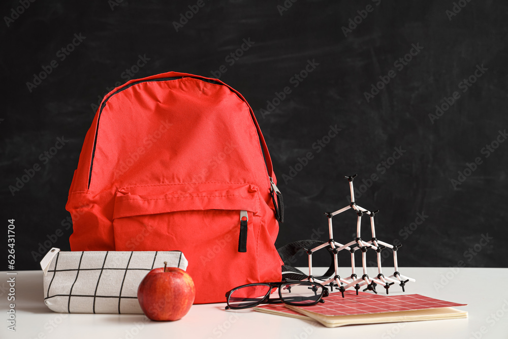 Red school backpack with stationery and molecular model on white table near black chalkboard