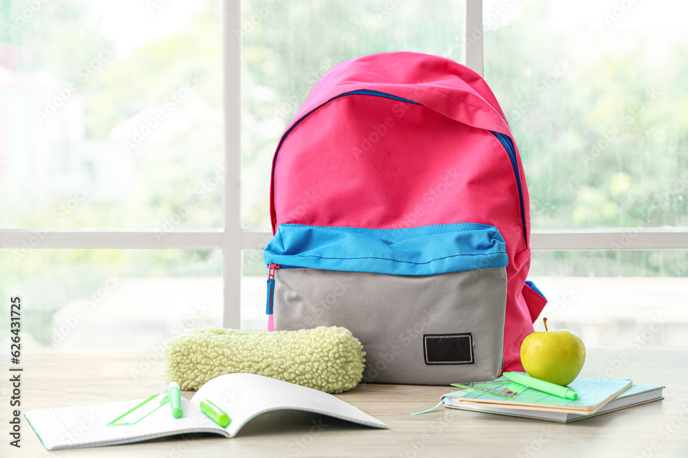 Colorful school backpack with stationery and fresh apple on wooden table near window in classroom