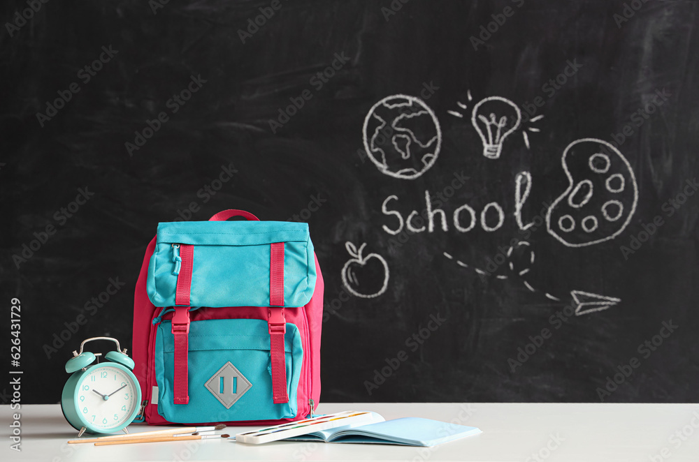 Colorful school backpack with stationery and alarm clock on white table near black chalkboard