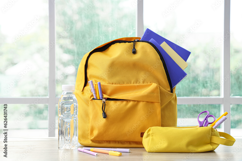 Yellow school backpack with stationery and bottle of water on wooden table near window in classroom