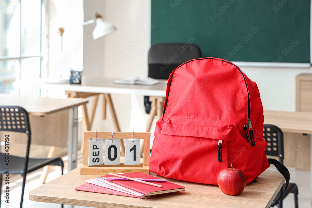 Red school backpack with stationery, calendar and fresh apple on desk in classroom