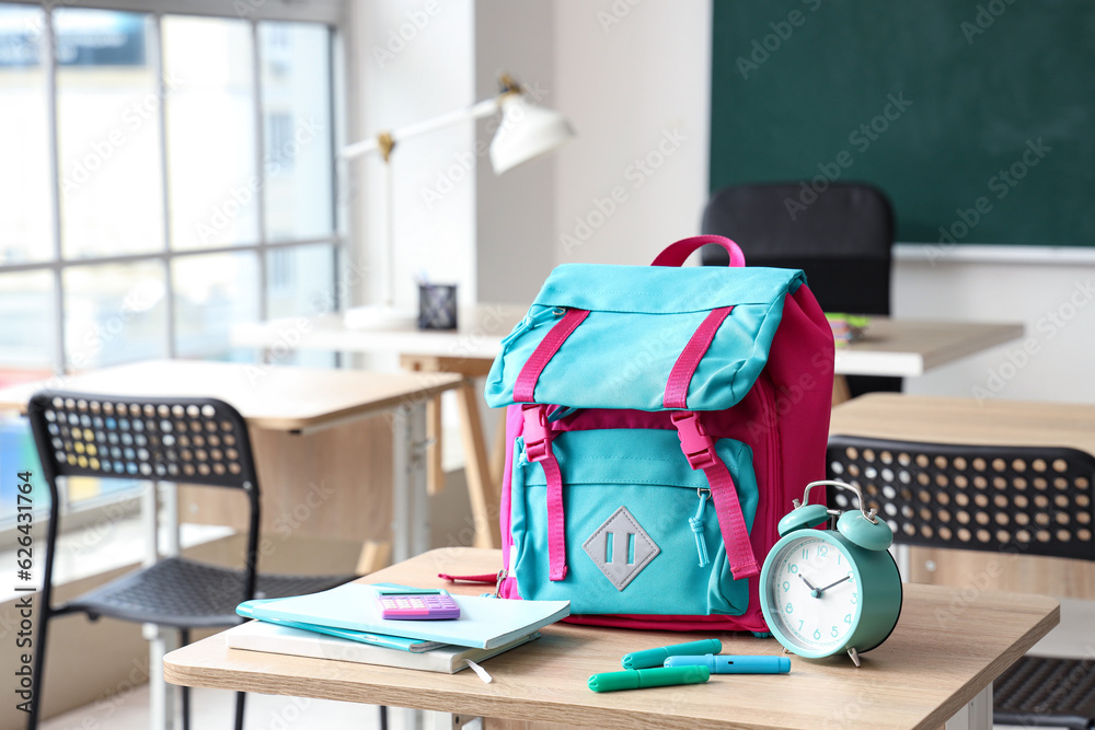 Colorful school backpack with stationery and alarm clock on desk in classroom