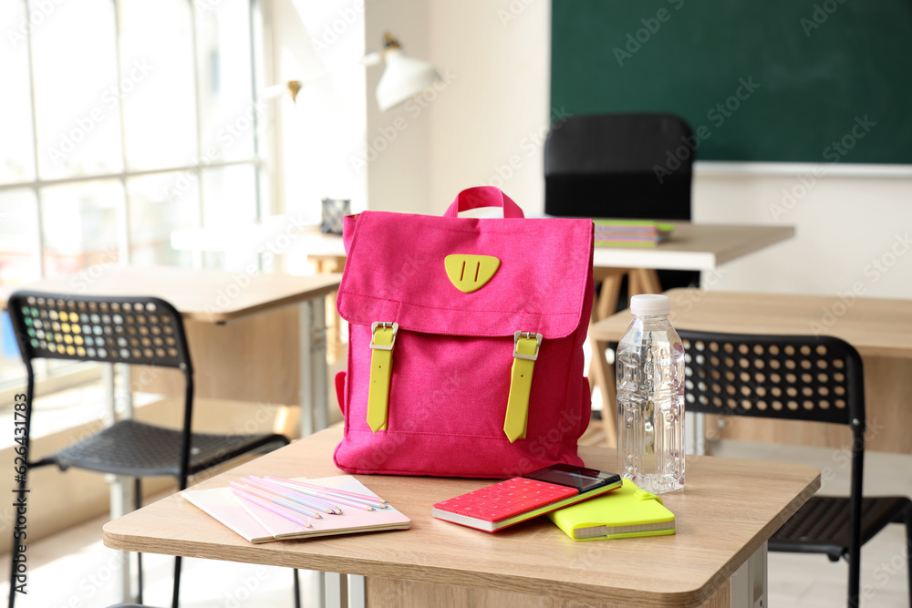 Pink school backpack with stationery and bottle of water on desk in classroom