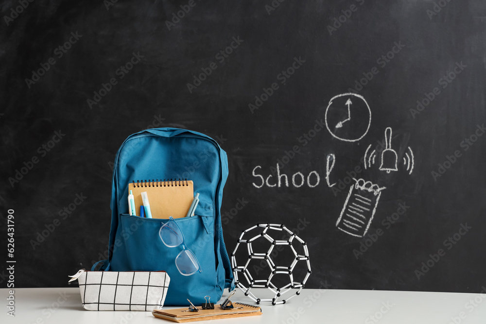 Blue school backpack with stationery and molecular model on white table near black chalkboard