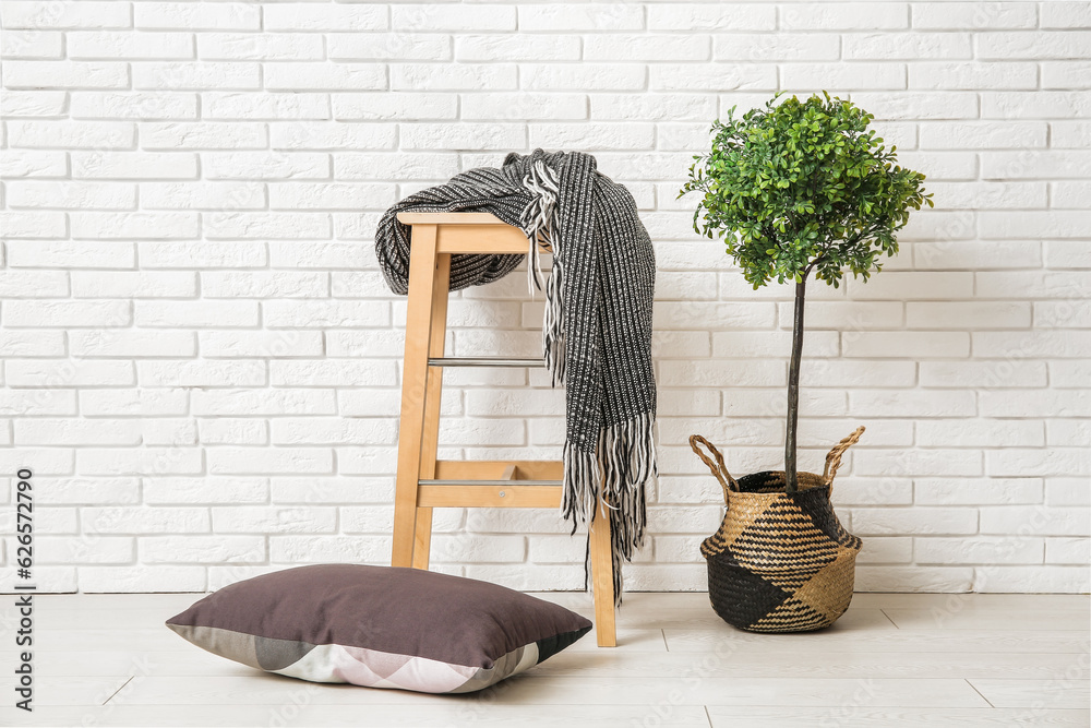 Stool with cozy blanket and cushions near white brick wall