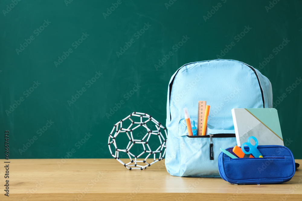 Backpack with school stationery and molecular model on table near green chalkboard