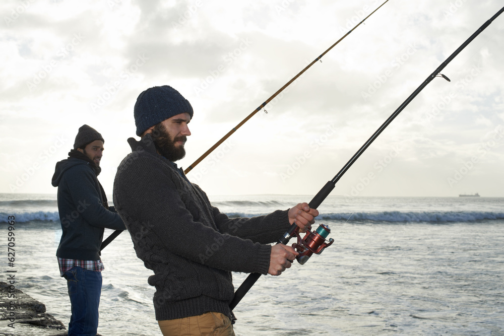 Secretly hoping hell hook the bigger fish...Shot of two young men fishing at the ocean in the early 