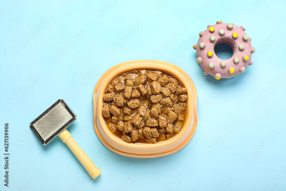 Bowl of wet pet food, toy and grooming brush on color background