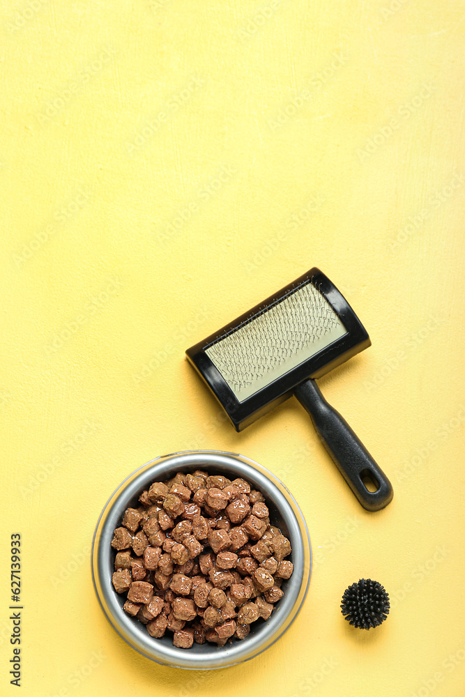 Bowl of wet pet food, toy and grooming brush on yellow background