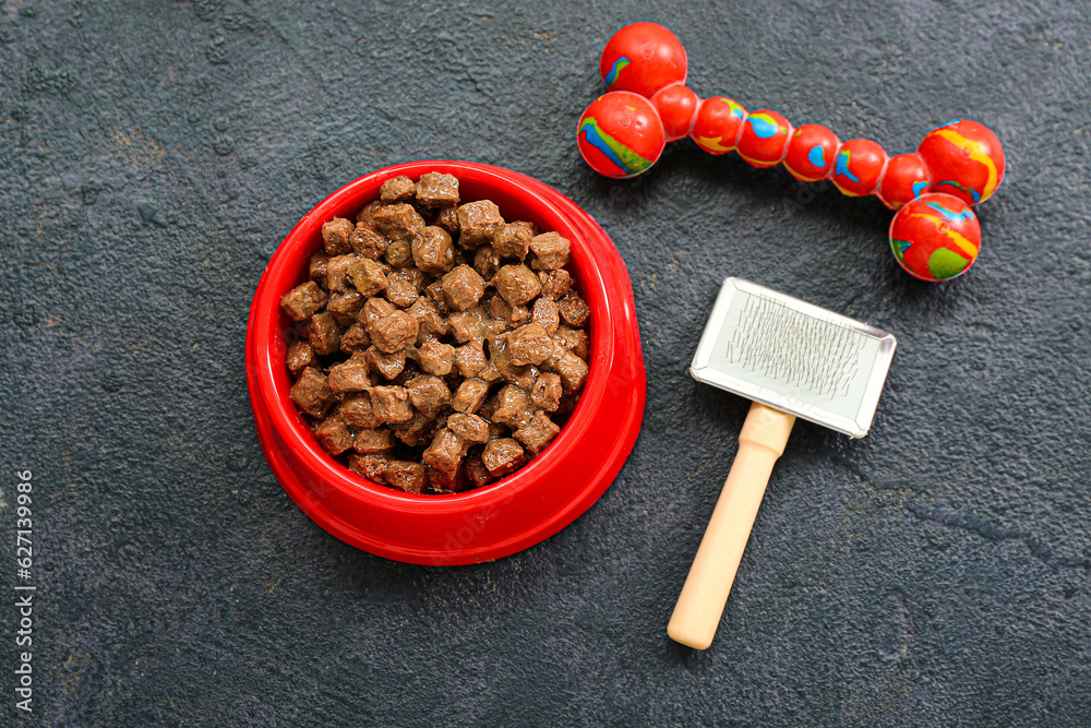 Bowl of wet pet food, rubber toy and grooming brush on dark background