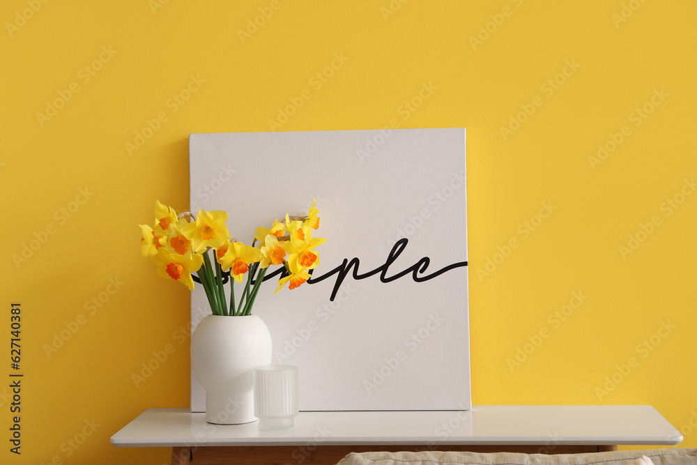 Vase with blooming narcissus flowers and poster on table near yellow wall