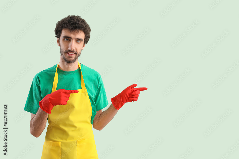 Young man in rubber gloves and apron pointing at something on green background