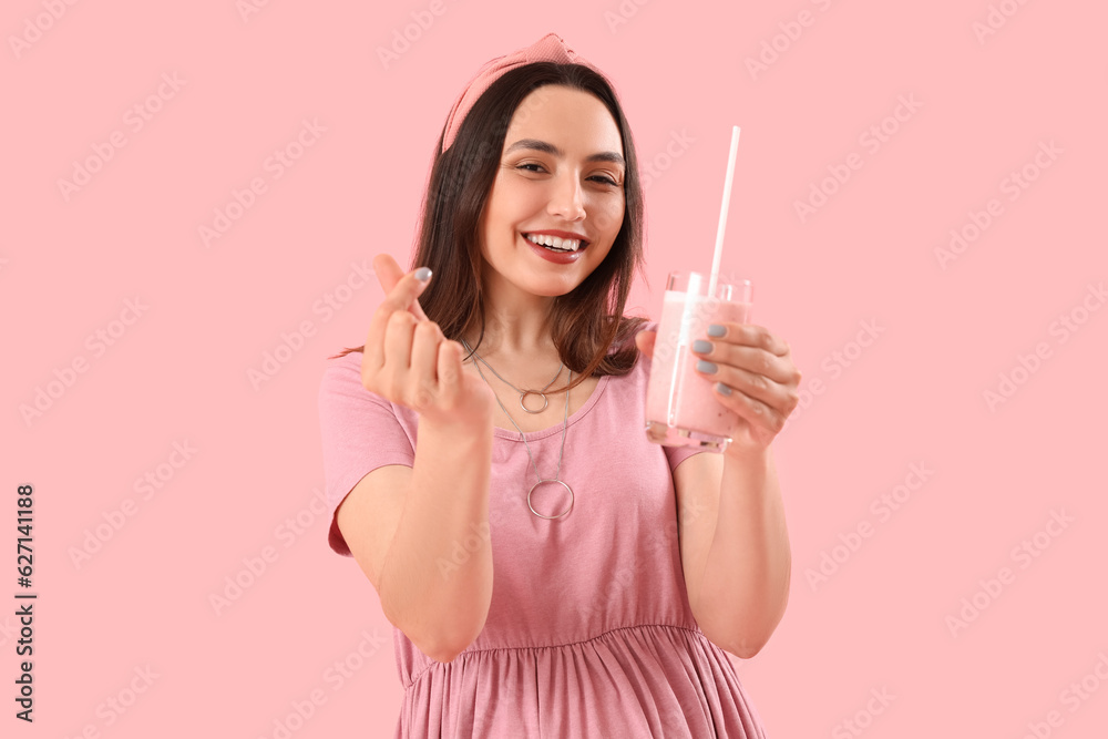 Young woman with glass of smoothie making heart on pink background