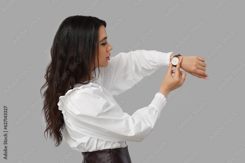 Beautiful young woman looking at wristwatch on grey background