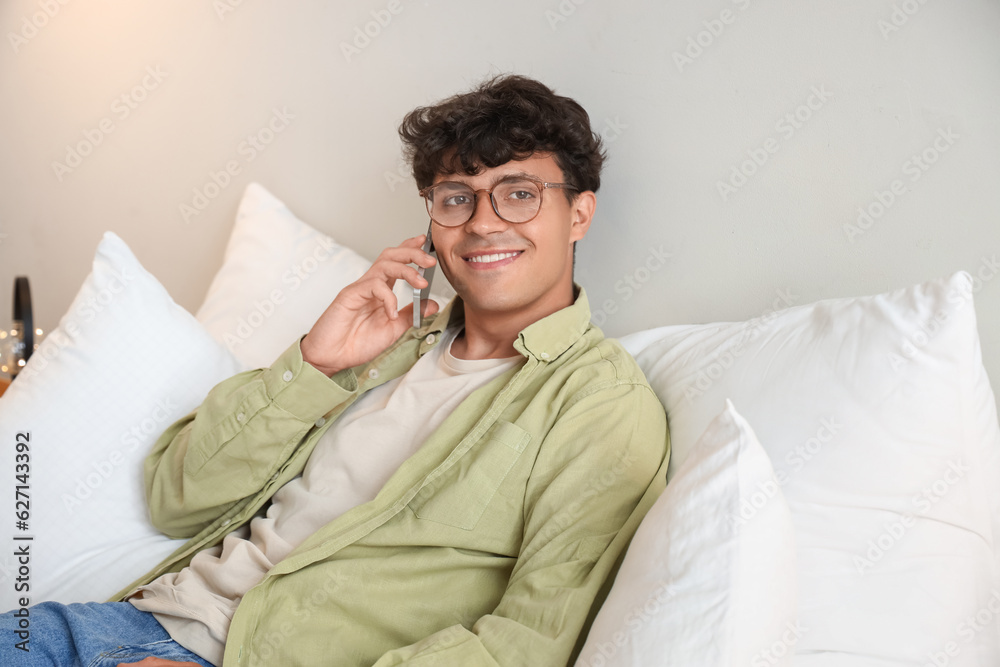 Young man with eyeglasses talking by mobile phone in bedroom