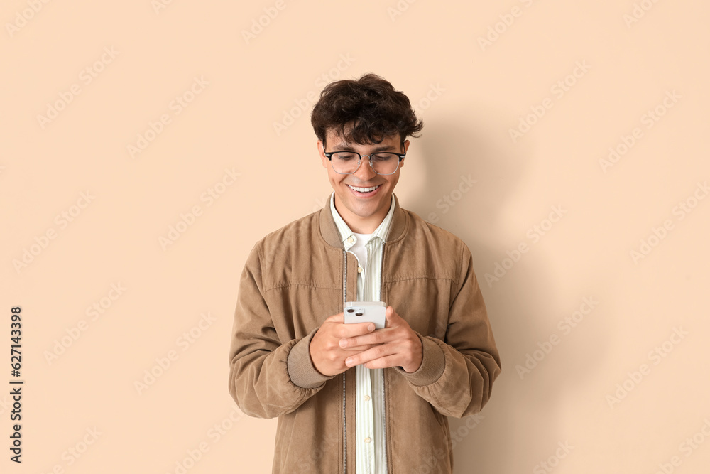 Young man in stylish eyeglasses using mobile phone on beige background