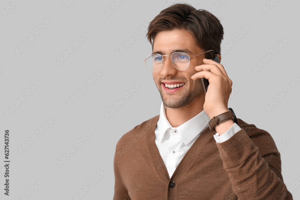 Stylish young handsome man in eyeglasses with wristwatch talking by mobile phone on grey background