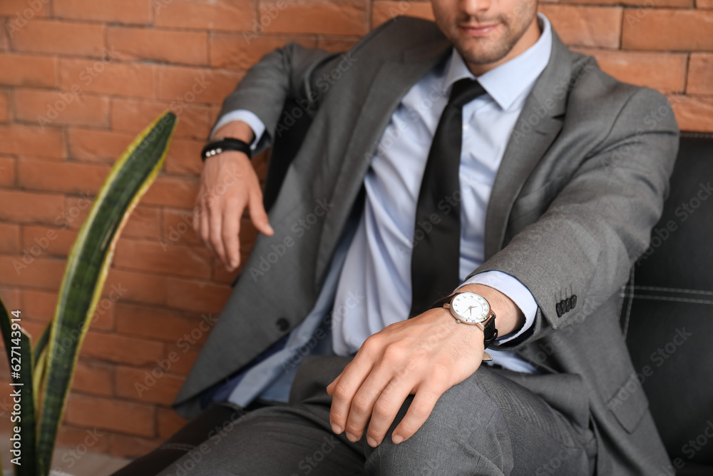 Stylish young man in suit with wristwatch sitting on leather sofa, closeup